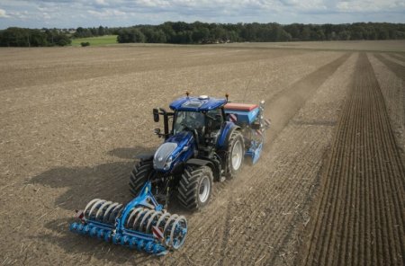 Traktor New Holland T7 se standardním rozvorem pracuje na poli s agregovaným secím strojem a zadním pěchem, který konsoliduje půdu za secími botkami.