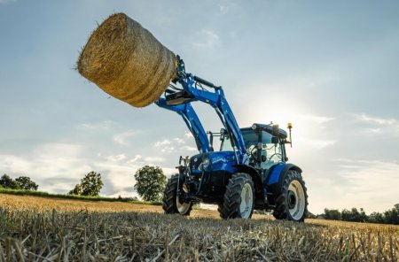Traktor New Holland T5S s čelním nakladačem zvedá kulatý balík slámy na strništi, kde vyniká jeho obratnost a síla.