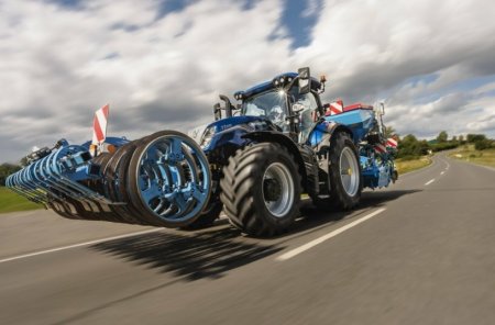 Traktor New Holland T7 se standardním rozvorem jede po silnici s agregovaným širokozáběrovým secím strojem a zadním půdním pěchem.