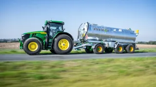 Traktor John Deere táhne stříbrnou cisternu Joskin Tetraliner RL po silnici.