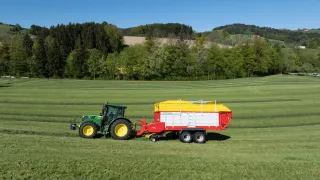 Zelený traktor s červeným sběracím vozem Pöttinger na louce