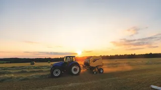 Modrý traktor s lisem při práci na poli za západu slunce