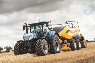 Modrý traktor New Holland táhne žlutý lis na hranaté balíky