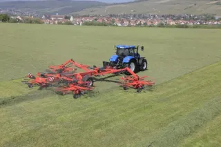 Modrý traktor táhne červený shrnovač Kuhn na louce