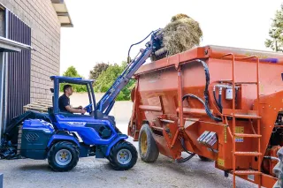 Modrý kloubový nakladač MultiOne nakládá seno do krmného vozu