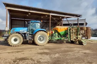 Modrý traktor New Holland s připojeným secím strojem Amazone před přístřeškem