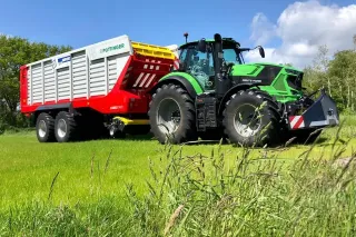 Zelený traktor táhne sběrací vůz Pöttinger Jumbo po louce