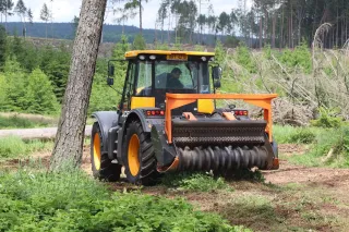 Žlutočerný traktor s oranžovým lesním mulčovačem pracuje na lesní cestě.