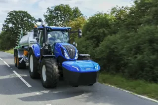 Modrý traktor New Holland táhne cisternový vůz po silnici.