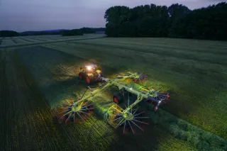 Čtyřrotorový shrnovač Claas Liner za traktorem pracuje na louce za šera.