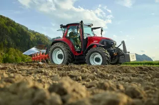 Červený traktor Lindner s pluhem pracuje na poli před horami.