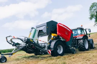 Červený traktor Massey Ferguson táhne lis na kulaté balíky po louce.