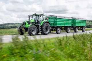 Zelený traktor Deutz-Fahr táhne dva zelené přívěsy po silnici.