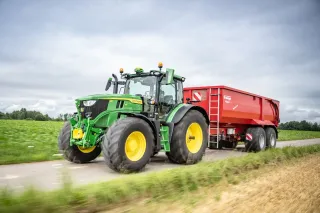 Zelený traktor John Deere 6R táhne červený přívěs.