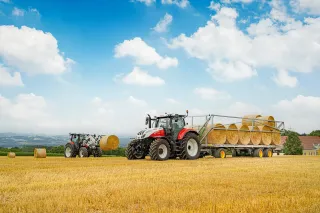 Traktor s přepravníkem naloženým kulatými balíky slámy na poli.