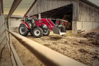 Červený traktor Case IH Farmall C s čelním nakladačem před stájí.