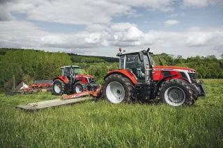Dva červené traktory Massey Ferguson se žací technikou na louce.