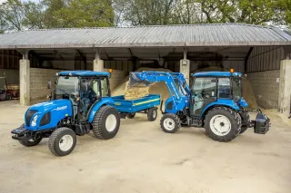 Dva modré traktory New Holland Boomer ve stodole; jeden nakládá materiál do přívěsu.