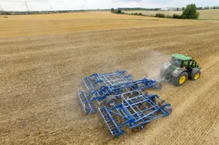 Traktor táhne široký modrý kypřič Köckerling Allrounder Flatline po poli.