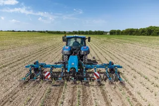 Modrý traktor New Holland s meziřádkovým kypřičem na poli.