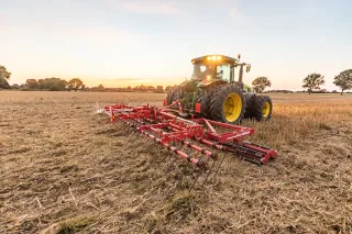 Traktor táhne červené nožové válce Horsch Cultro TC po poli při západu slunce.