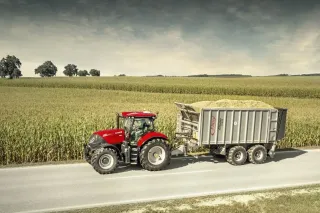 Červený traktor Case IH Puma veze přívěs s nákladem po silnici u pole.