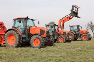 Tři oranžové traktory Kubota na louce, prostřední s čelním nakladačem.