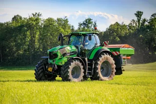 Zelený traktor Deutz-Fahr s rozmetadlem na travnatém poli.