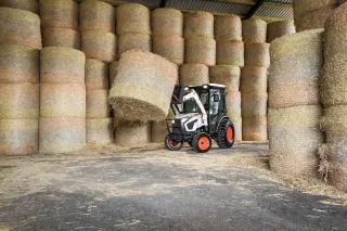 Bílý komunální traktor Bobcat zvedá balík slámy ve skladu.