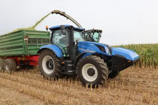 Modrý traktor New Holland při odvozu zelené hmoty od řezačky.