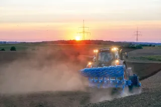 Traktor s modrým pluhem Lemken Diamant 16 při orbě za západu slunce.