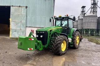 Zelený traktor John Deere před hospodářskou budovou.
