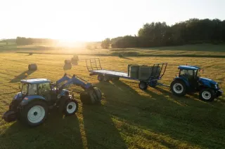 Dva modré traktory New Holland při manipulaci s balíky na louce.