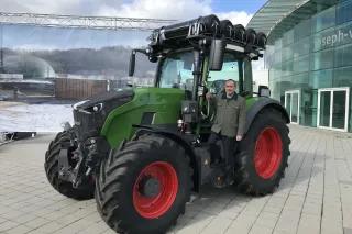 Zelený traktor Fendt s mužem před budovou.