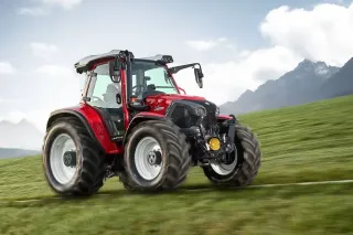 Červený traktor Lindner na travnatém svahu před horskou krajinou.