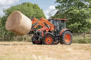 Oranžový traktor Kubota zvedá balík sena.