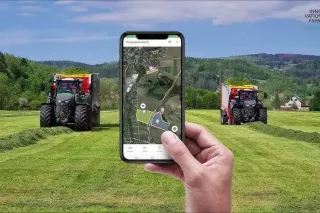 Mobilní telefon s aplikací Harvest Assist před traktory na louce.