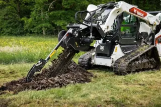 Pásový nakladač Bobcat s rýhovačem hloubí rýhu v travnatém terénu.