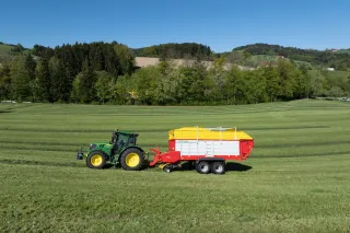 Zelený traktor s červeným sběracím vozem Pöttinger na louce