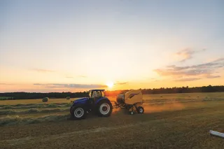 Modrý traktor s lisem při práci na poli za západu slunce