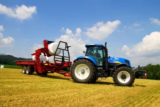 Modrý traktor New Holland veze na přívěsu Anderson bílé balíky.
