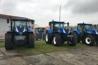 Tři modré traktory New Holland na venkovní výstavě.