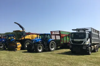 Modrý traktor New Holland vedle řezačky a nákladního vozu.
