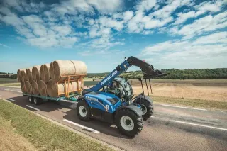 Modrý manipulátor New Holland převážející kulaté balíky na přívěs