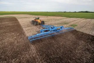 Traktor táhne široký modrý stroj na strništi