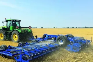 Zelený traktor táhnoucí široký modrý kultivátor po poli