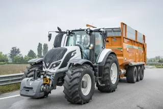 Bílý traktor Valtra táhne oranžový třínápravový přívěs