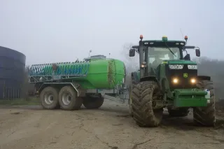 Zelený traktor John Deere táhne zelenou dvounápravovou cisternu Bauer na dvoře.