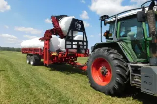 Traktor Fendt táhne červený přepravník Anderson s fóliovanými balíky.