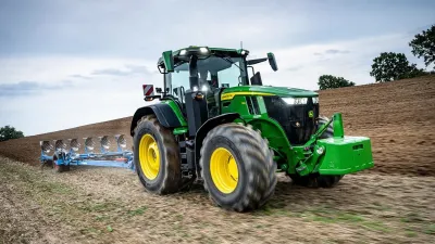 Zelený traktor John Deere s pluhem na poli.
